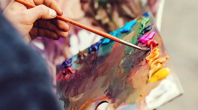 Close up of colourful paint-covered artist's palette with a hand holding a small paintbrush