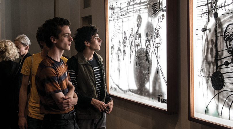 Two people looking at two large black and white brightly lit drawings in a dark gallery.