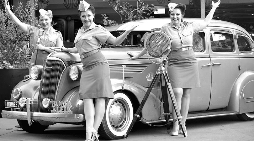 black and white image of 3 ladies in old army outfits next to old car with lights and microphone