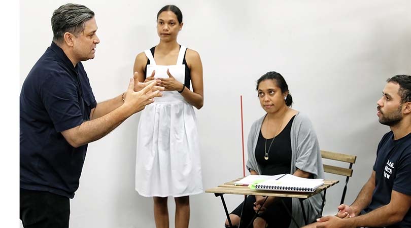 four indigenous performers, one man and one women standing, one man and one woman sitting with desk and books in front of her, standing man has hands in air describing something