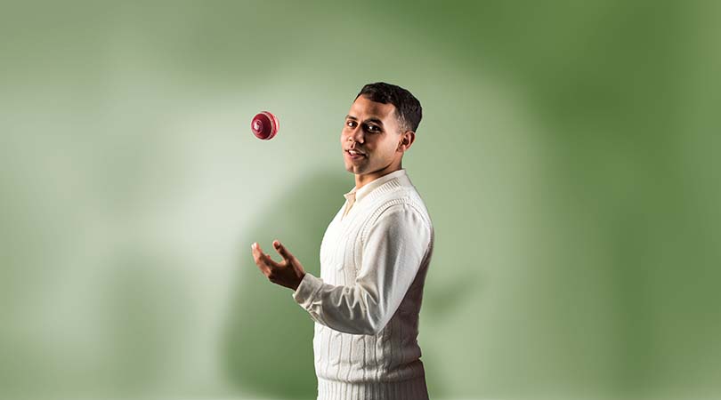 man standing in front of green backdrop wearing cricket uniform, throwing a red cricket ball up in the air