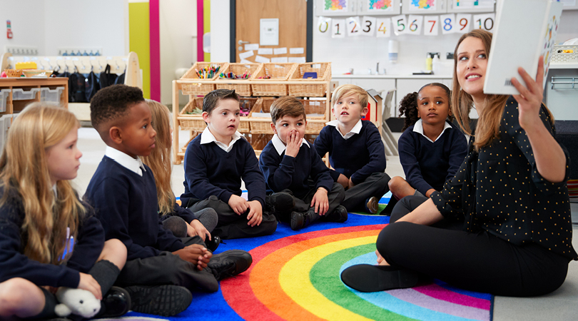seven young children in blue and grey school uniform, sitting on rainbow rug being read a story by young female teacher in classroom