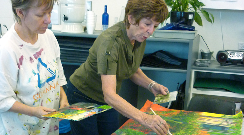 two older ladies, one leaning over and painting, one holding the paint board next to her