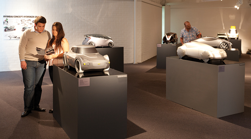 silver model cars on large grey plinths with a couple standing looking at a program and another man behind looking at one of the cars