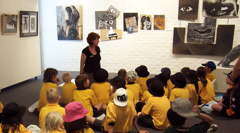 group of school kids in yellow uniform sitting on floor of art gallery in front of teacher with art work on walls behind