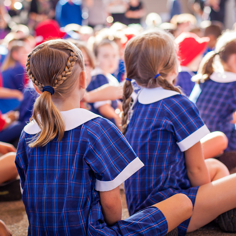 backs of school children in blue check uniform sitting on floor