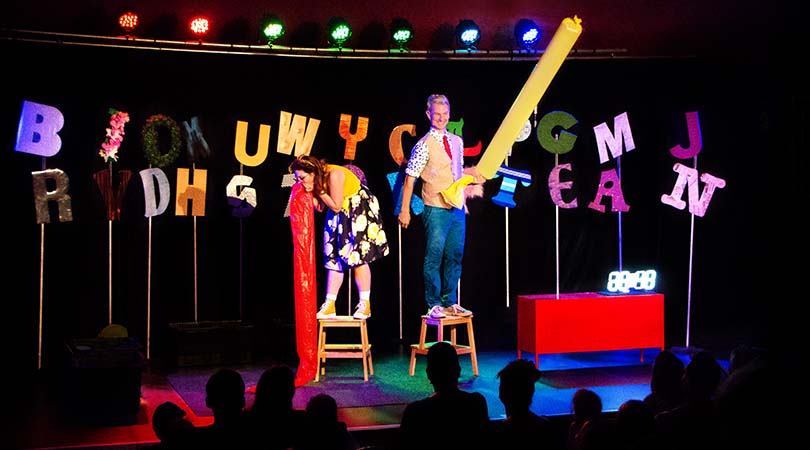 lady standing on stool leaning over blowing up a large red balloon. Man standing on stool next to her holding up a large yellow inflated balloon. Letters on sticks and stage lights behind them.