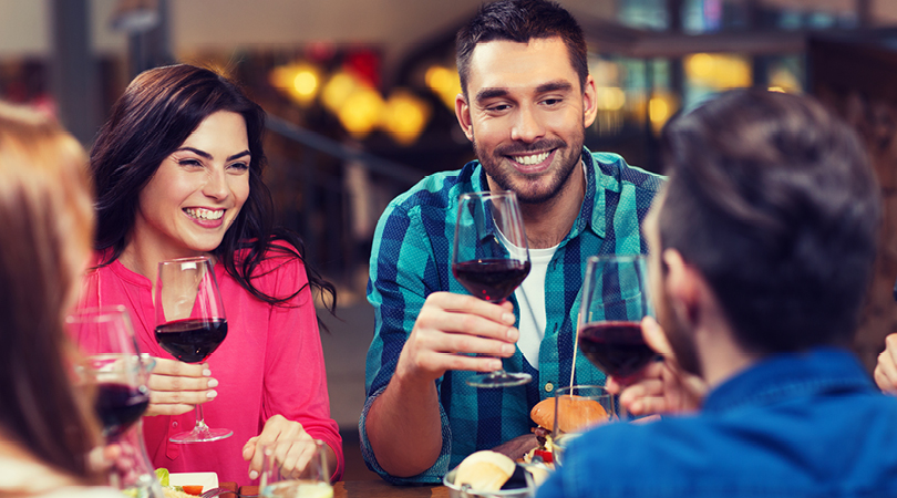 man and woman smiling drinking red wine talking to another couple in front of them with the backs of their heads in shot