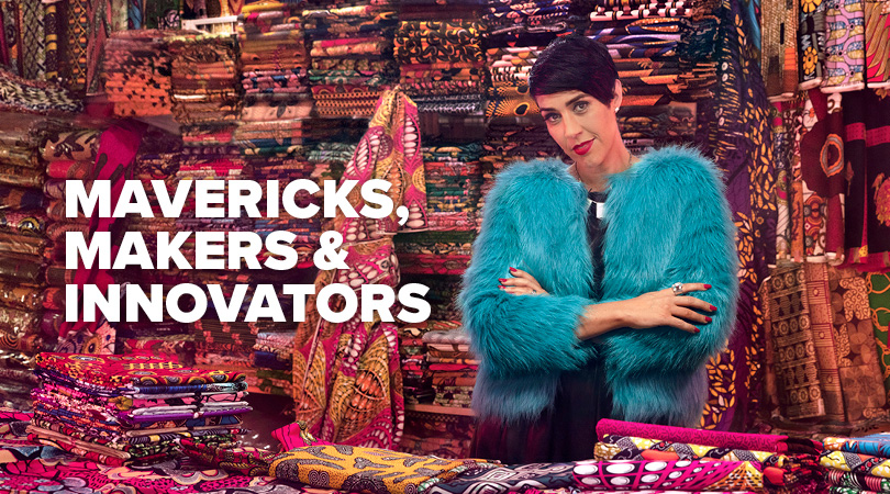 Mavericks, Makers & Innovators, lady is fluffy blue jacket with arms folded, piles and piles of colourful fabric behind her