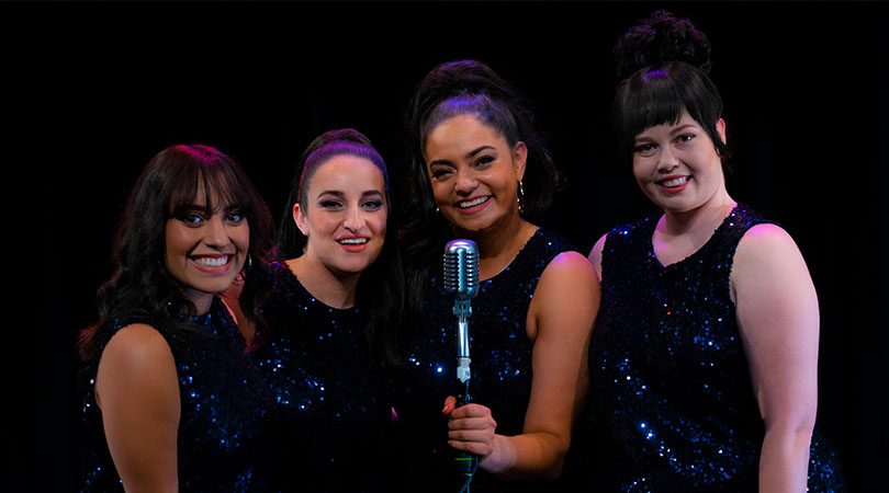 four ladies in blue sequin dresses singing into old style microphone