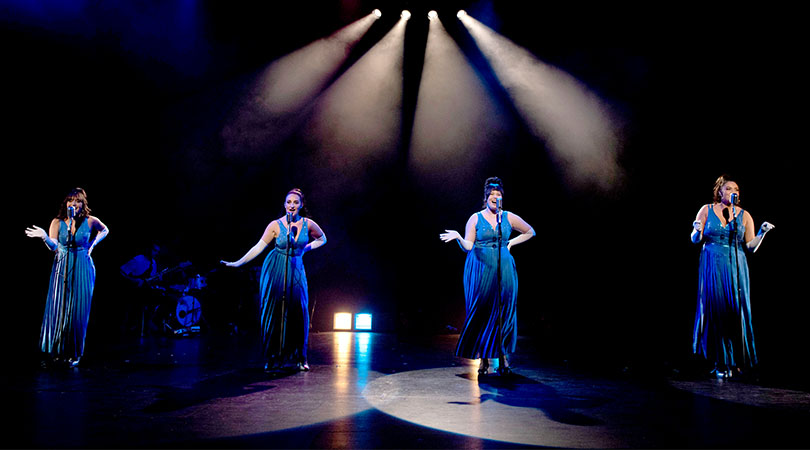 four ladies on stage singing into microphones wearing long blue dresses with stage lights above