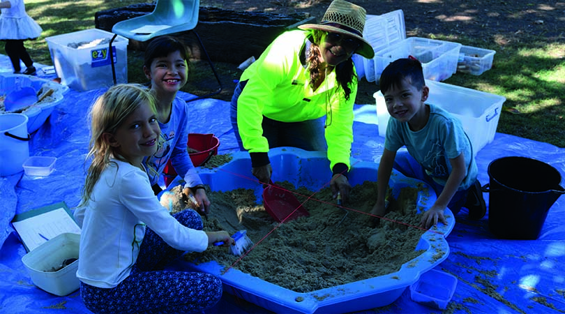 Three children and a volunteer are doing an archaeological dig in a sandpit.