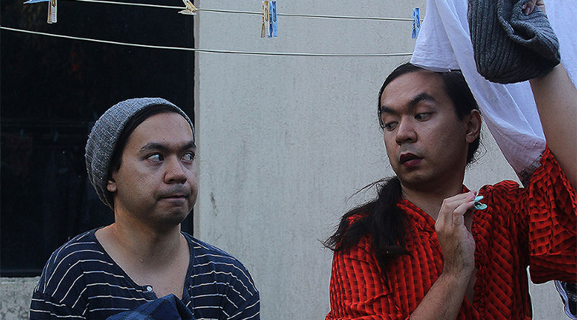 A man in a beanie stares cautiously towards a man in a red dress hanging up washing