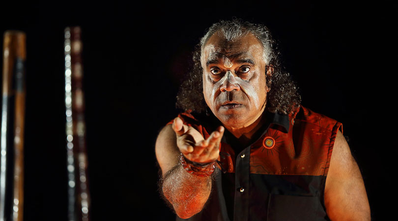 Australian indigenous man with long curly hair pulled back, wearing tribal face paint, holding his hand out in front of his face with a black background