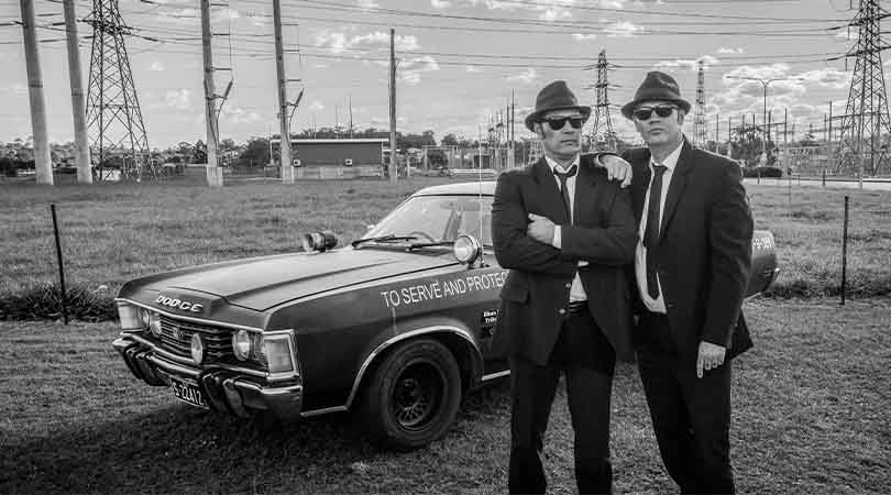 black and white image of two men in suits, with hats and sunglasses standing next to an old police car