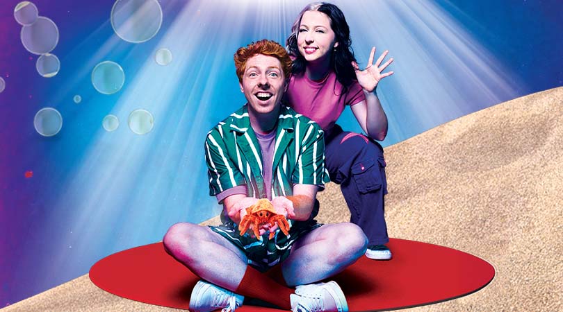 Two people on sand under blue light beams; one sits cross-legged on a red mat holding a starfish, the other kneels behind.