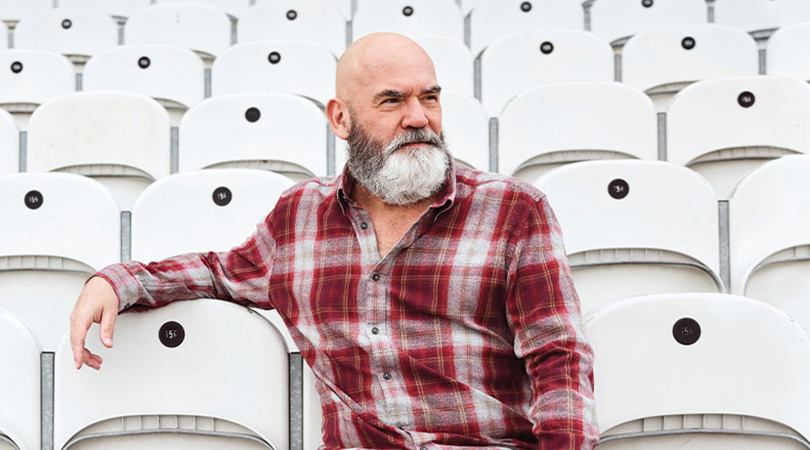 A bearded adult wearing a red plaid shirt sits alone among rows of light grey stadium-style seats, resting one arm across the back of a chair and looking to the side.